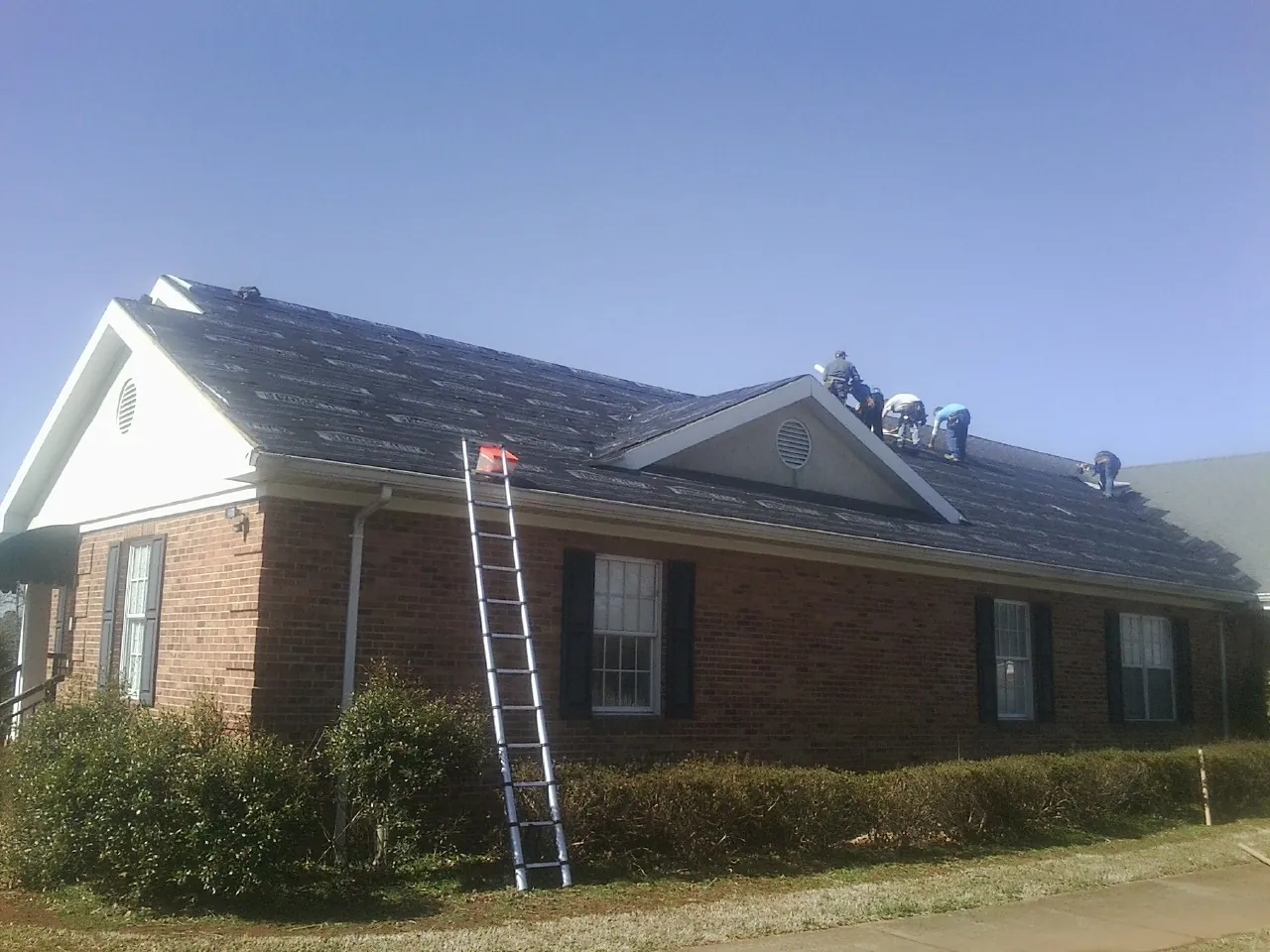 Crew tearing shingles off a brick ranch roof with a ladder set — Metal Roof Repair work by our crew in Fort Atkinson
