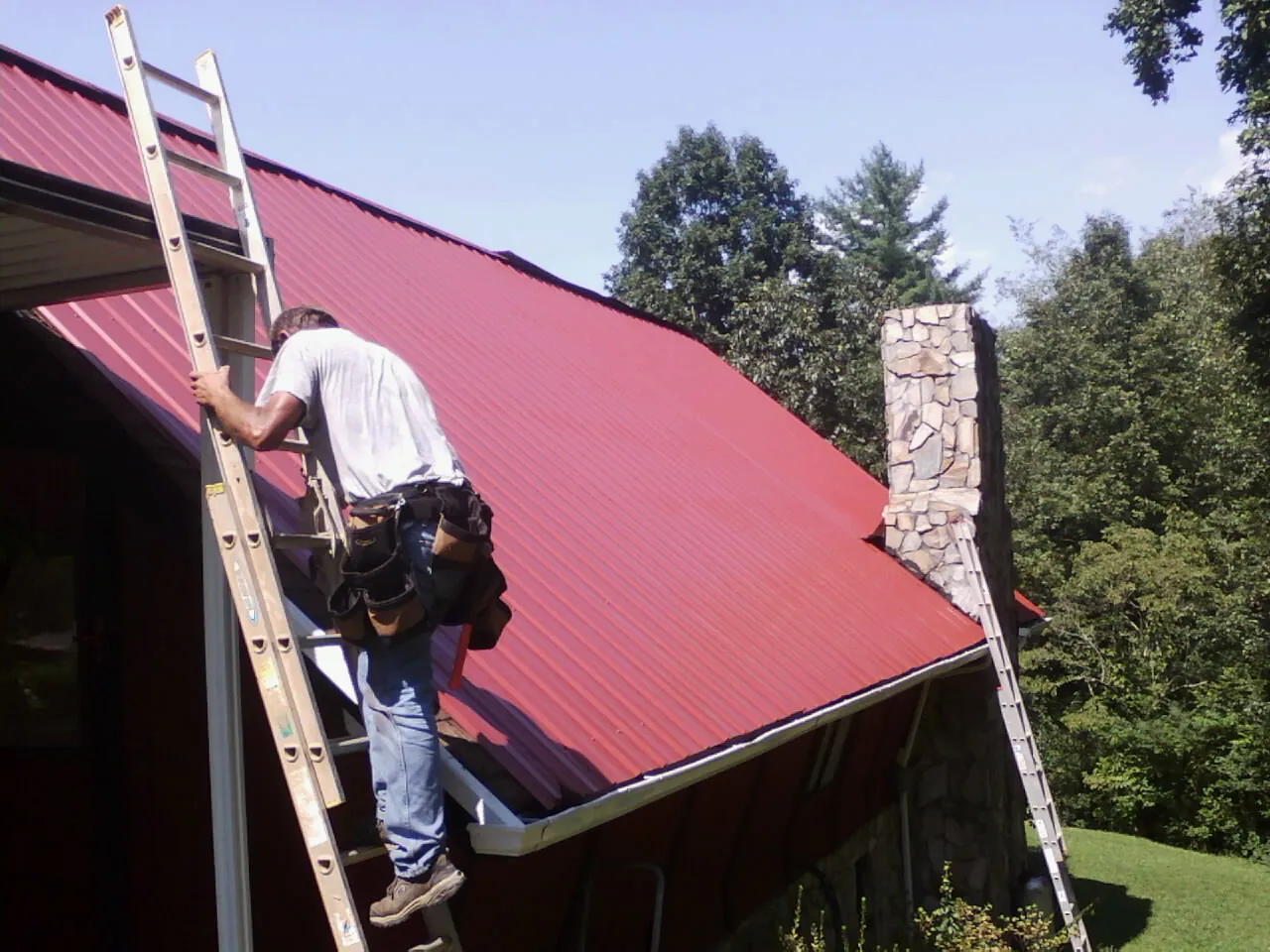 Roofer on a ladder beside a red metal roof and stone chimney — Soffit & Fascia Repair by Roofing Brothers in Fort Atkinson